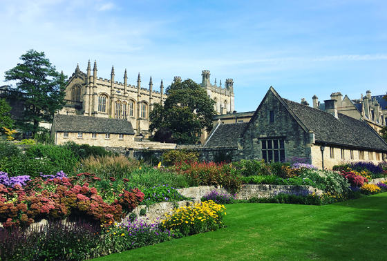 Lush green lawn and flowering gardens in the foreground with an English church and stone buildings in the background
