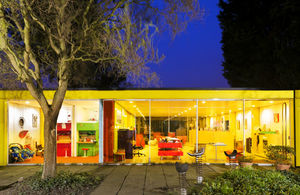 A modern, single-story house with large glass windows showcasing a brightly lit interior with colorful furniture and a tree and garden visible outside at dusk