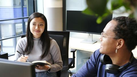 Two coworkers engaged in a discussion in an office setting with computers 