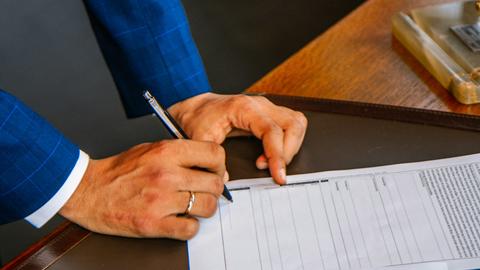 A person in a blue suit signing a document on a dark table