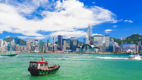 Hong Kong harbor and skyline with numerous high-rise buildings under a partly cloudy blue sky and several boats in the water