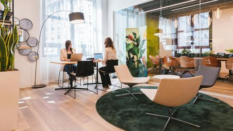 An office lounge featuring two women engaging in conversation and seated at a small table near large windows and a conference room