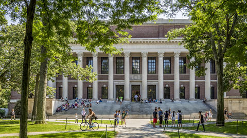 A large, classical-style academic building with columns and wide steps, surrounded by blue sky, trees, a grassy lawn, and people