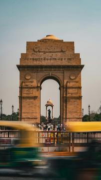 India Gate, a large sandstone arch in New Delhi, with people gathered at its base and blurred yellow and green auto-rickshaws passing in the foreground