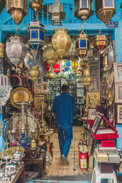 A man in a blue jacket walks through a market stall filled with ornate brass lanterns, hookahs, framed artwork, and Middle Eastern and North African crafts