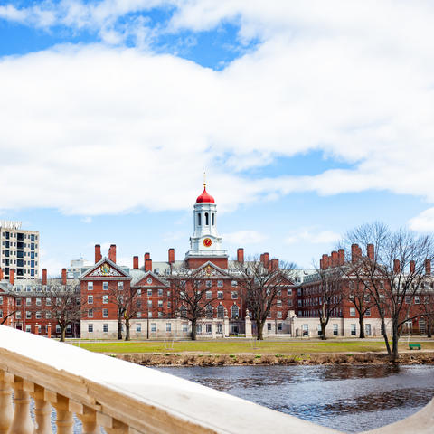A stone bridge with lampposts crossing a river with red-brick academic buildings under a partly cloudy blue sky
