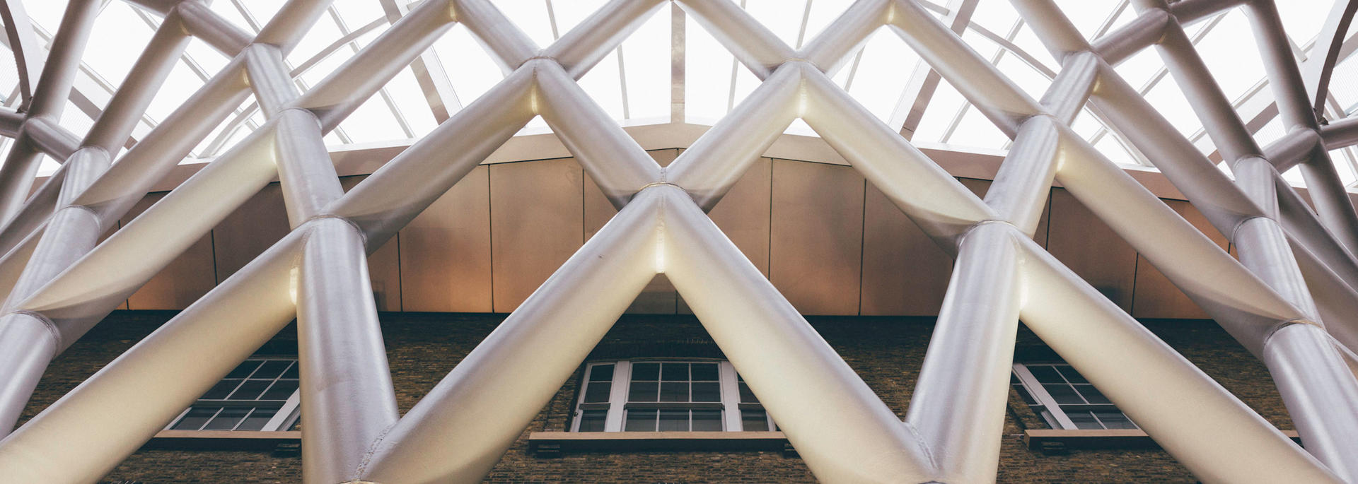 An upward view of a brick building facade framed by a geometric metal lattice structure with large, intersecting diagonal beams