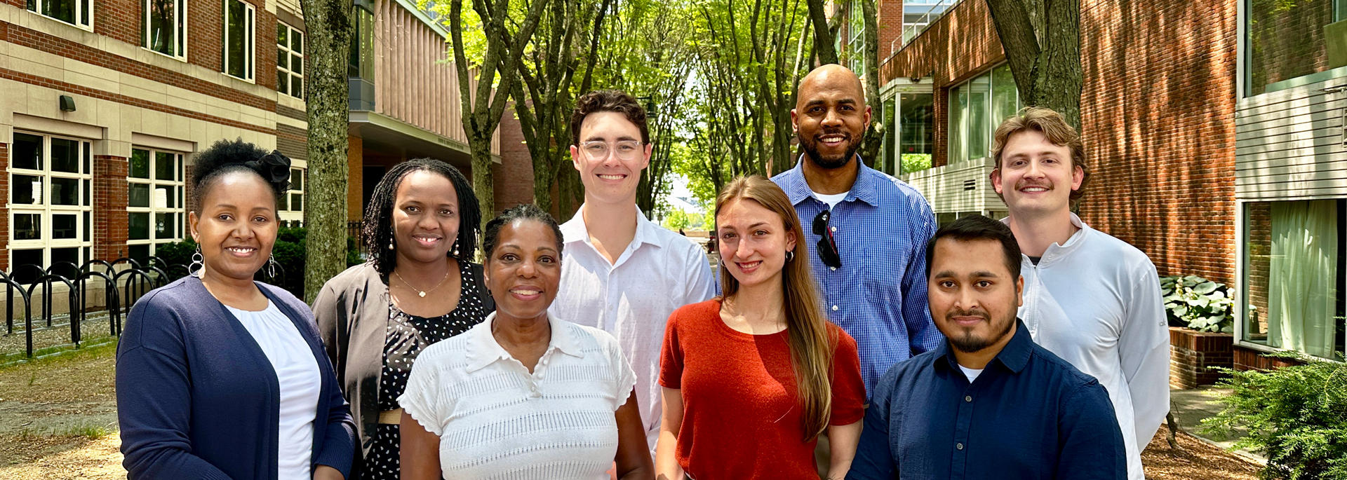 A group of eight people wearing business casual attire stand together on a walkway between two brick buildings with trees overhead on a sunny day