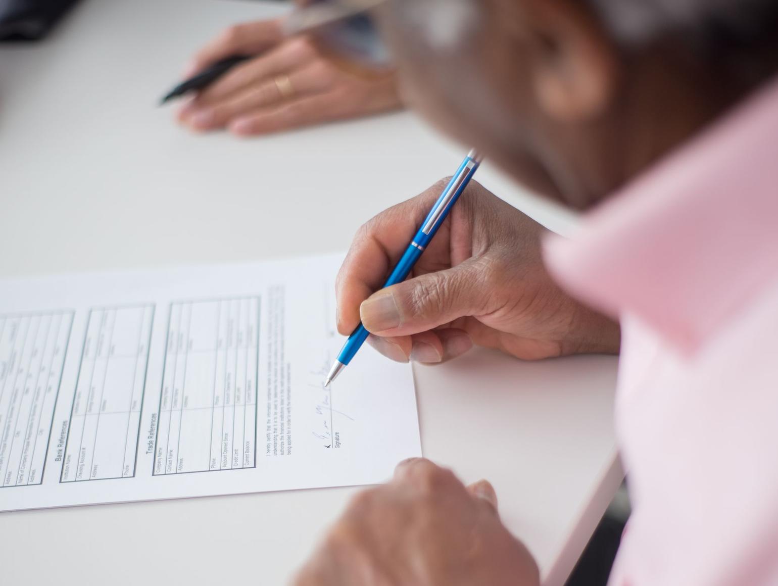 A close-up of a person sitting at a desk and signing a document with a blue pen