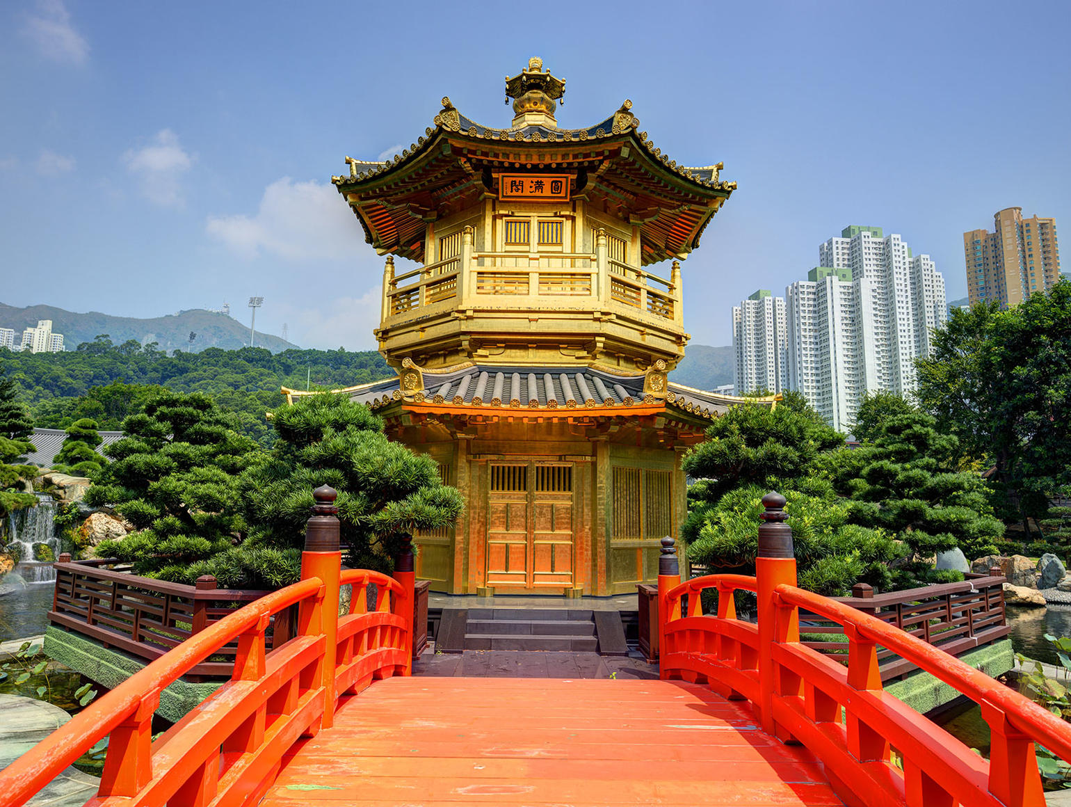 Gold Pavilion of Perfection in Hong Kong situated in a garden with a bright red arched bridge leading to its entrance and high-rise buildings in the background