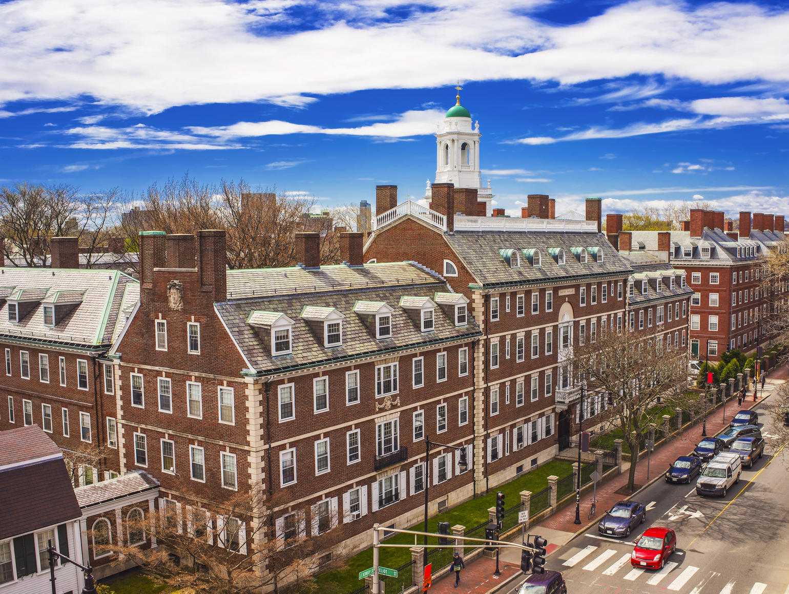 Red brick buildings and a bell tower under a partly cloudy blue sky next to a street with cars, pedestrians, and trees 