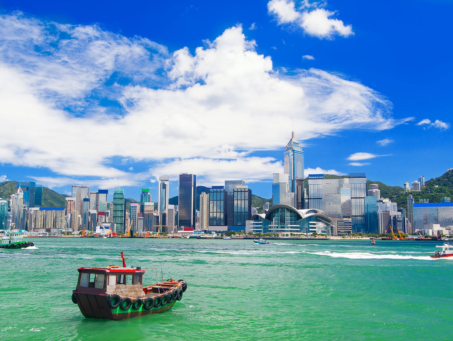 Hong Kong harbor and skyline with numerous high-rise buildings under a partly cloudy blue sky and several boats in the water