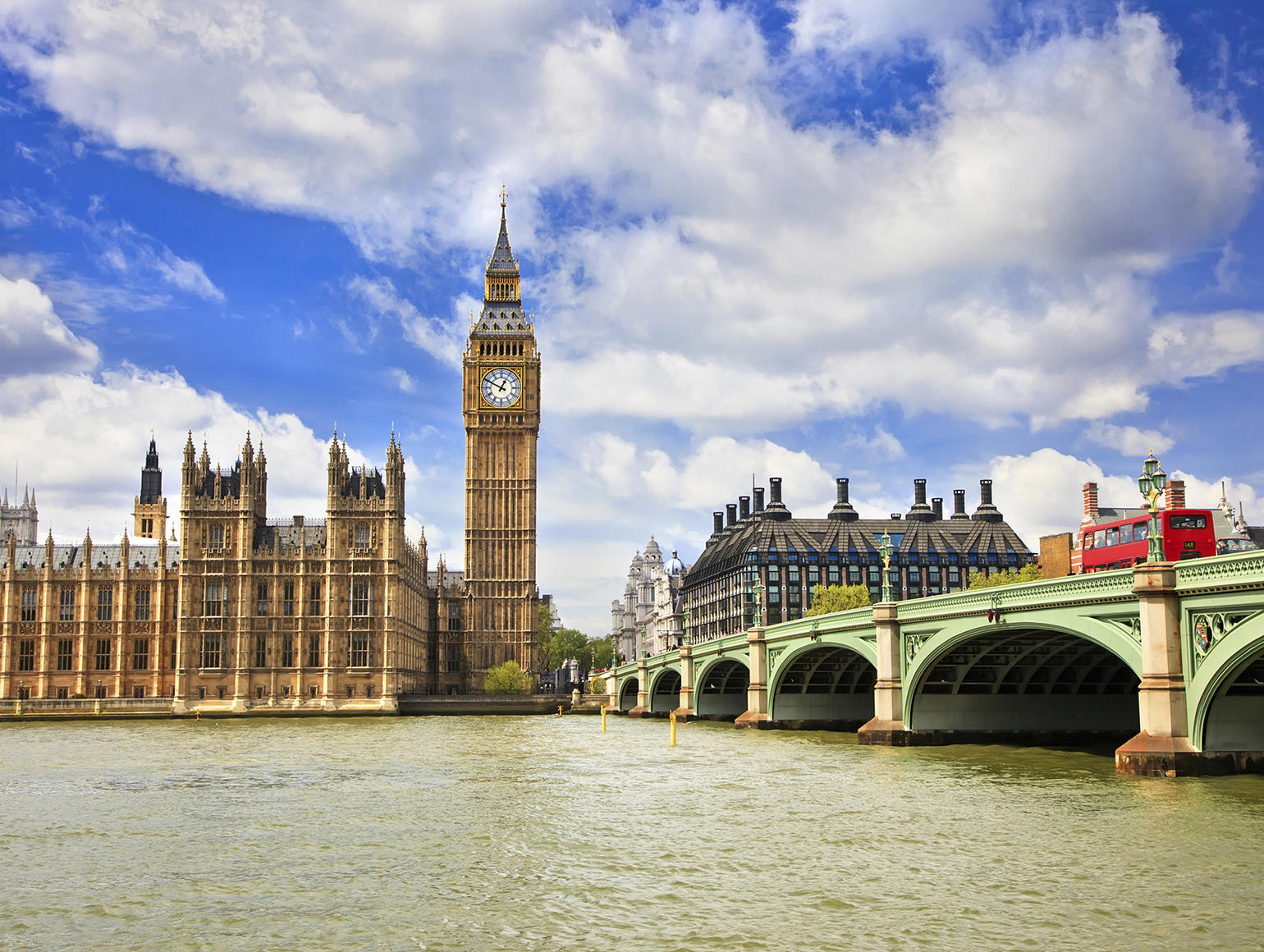 Palace of Westminster and Big Ben alongside Westminster Bridge with a red double-decker bus crossing the River Thames under a bright blue sky