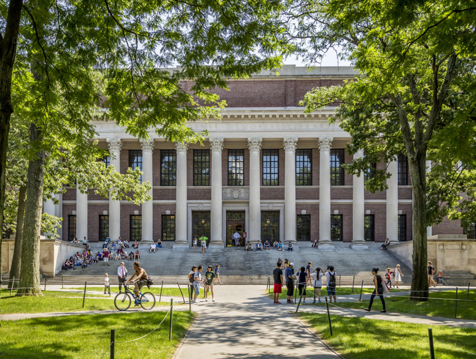 A large, classical-style academic building with columns and wide steps, surrounded by blue sky, trees, a grassy lawn, and people