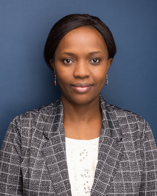 A woman with dark brown eyes and dark hair wearing a gray checkered blazer and white blouse, set against a solid dark blue background