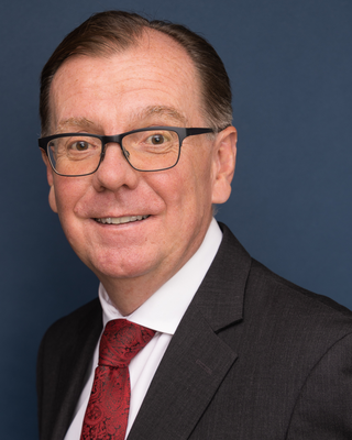 Man in glasses, dark suit, and red patterned tie smiling against blue