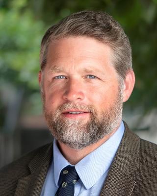 A man with blue eyes and a beard in brown blazer and blue shirt posing outdoors with leafy background