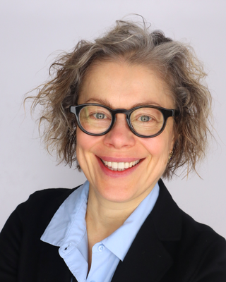 A smiling woman with short, curly light hair wearing black-rimmed glasses, a light blue collared shirt, and a black blazer