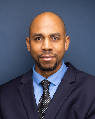 A man with dark brown eyes, shaved head, and trimmed beard wearing a dark suit, light blue shirt, and black polka dot tie against a dark blue background