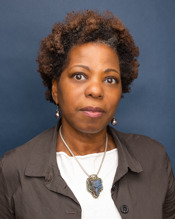 A woman with dark brown eyes and short curly hair wearing a dark jacket, white shirt, and a distinctive pendant necklace against a solid blue background