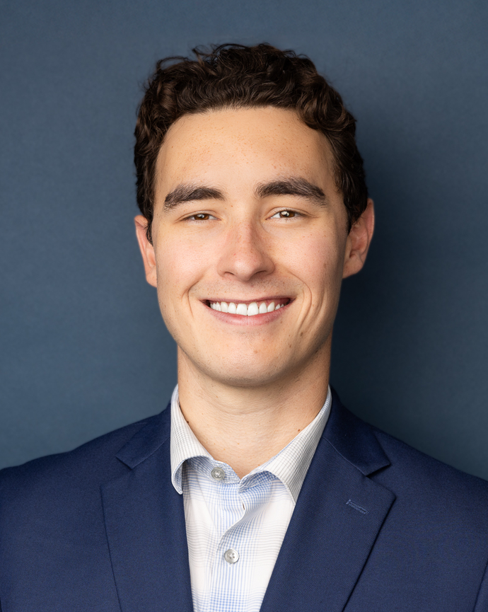 Man with dark curly hair in a navy blazer and light dress shirt smiling against a dark blue backdrop.