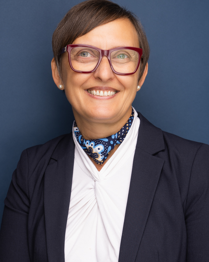 A smiling woman wearing large red glasses, a navy blazer, white blouse, and a patterned blue neck scarf against a solid dark blue background