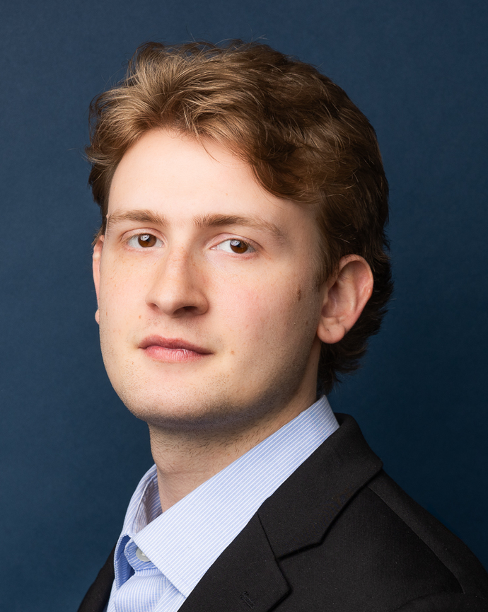 Man with blonde hair and brown eyes in a black blazer and blue shirt looking at camera before a blue backdrop