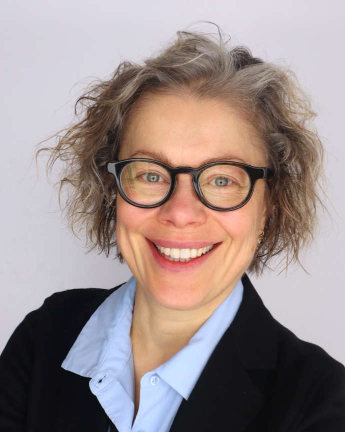 A smiling woman with short, curly light hair wearing black-rimmed glasses, a light blue collared shirt, and a black blazer