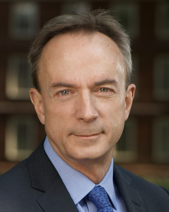 A man with short brown hair and light green eyes wearing a dark suit jacket, light blue shirt, and blue patterned tie
