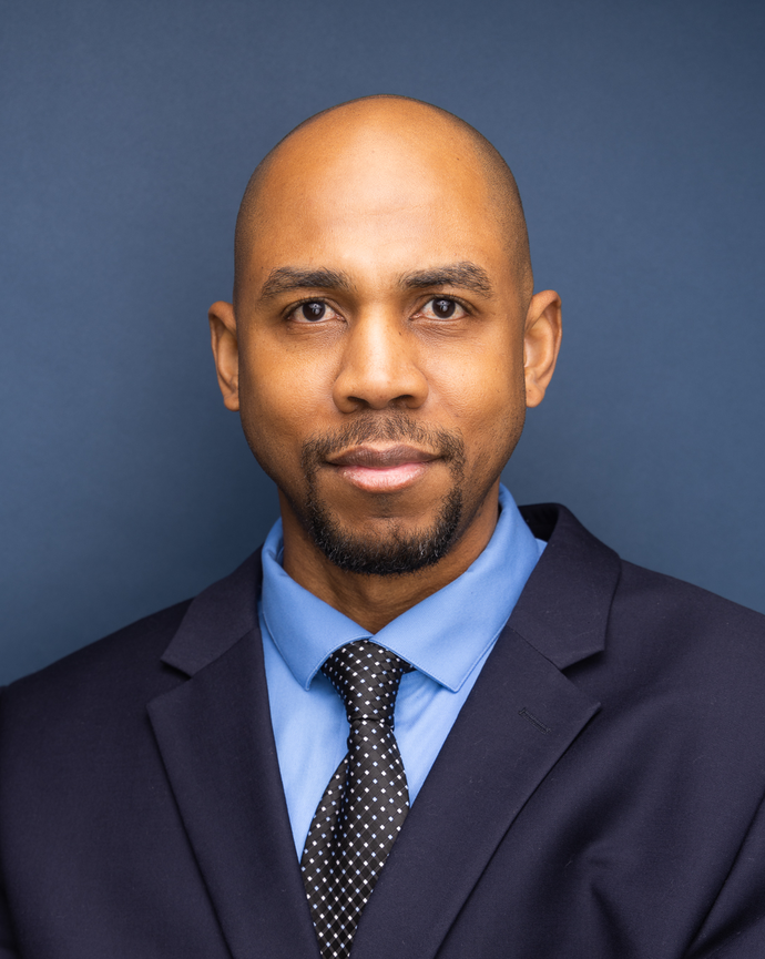 A man with dark brown eyes, shaved head, and trimmed beard wearing a dark suit, light blue shirt, and black polka dot tie against a dark blue background