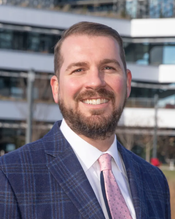 A smiling man with short brown hair and a beard, wearing a blue plaid suit jacket, white shirt, and pink tie
